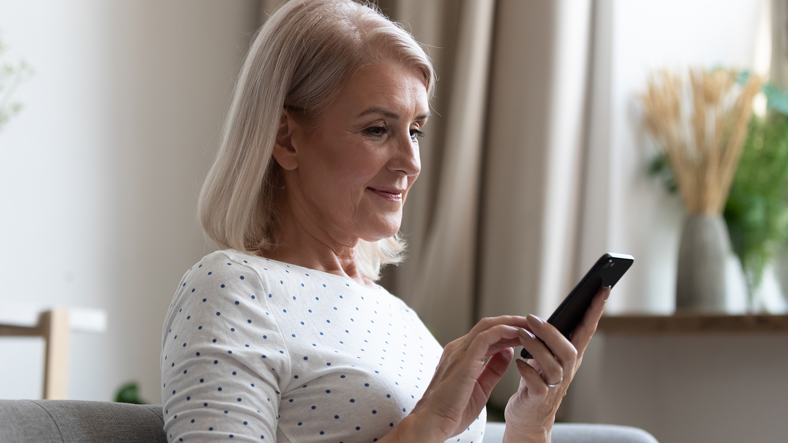 Happy middle aged mature woman enjoying using mobile apps texting typing messages sit on sofa, smiling old adult lady holding smartphone looking at cellphone screen browsing social media at home
