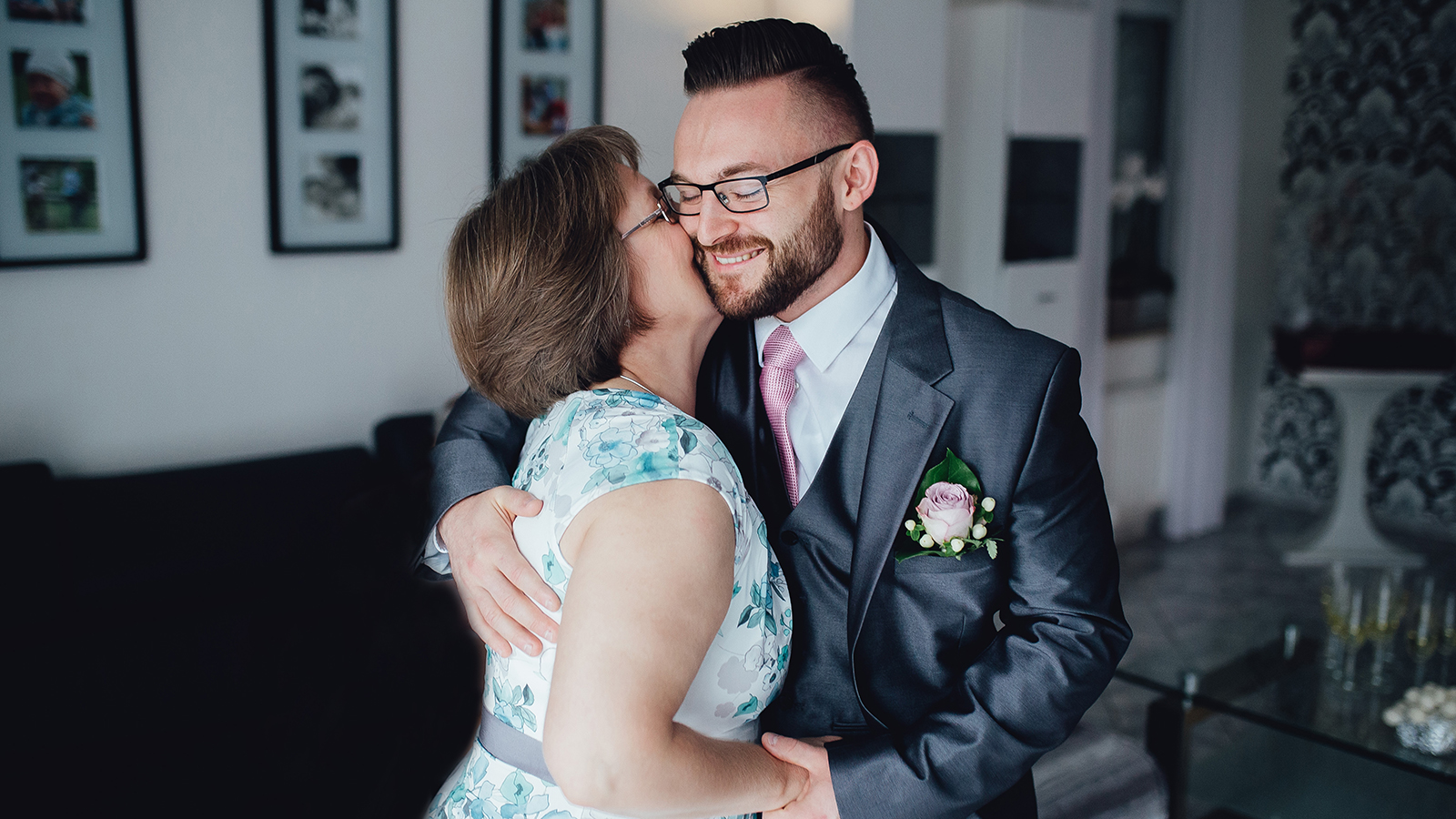 Portrait of a handsome groom in a grey suit with a tie with his mother. They are standing in the dining room, embracing each other and mother kissing her son and they are smiling.