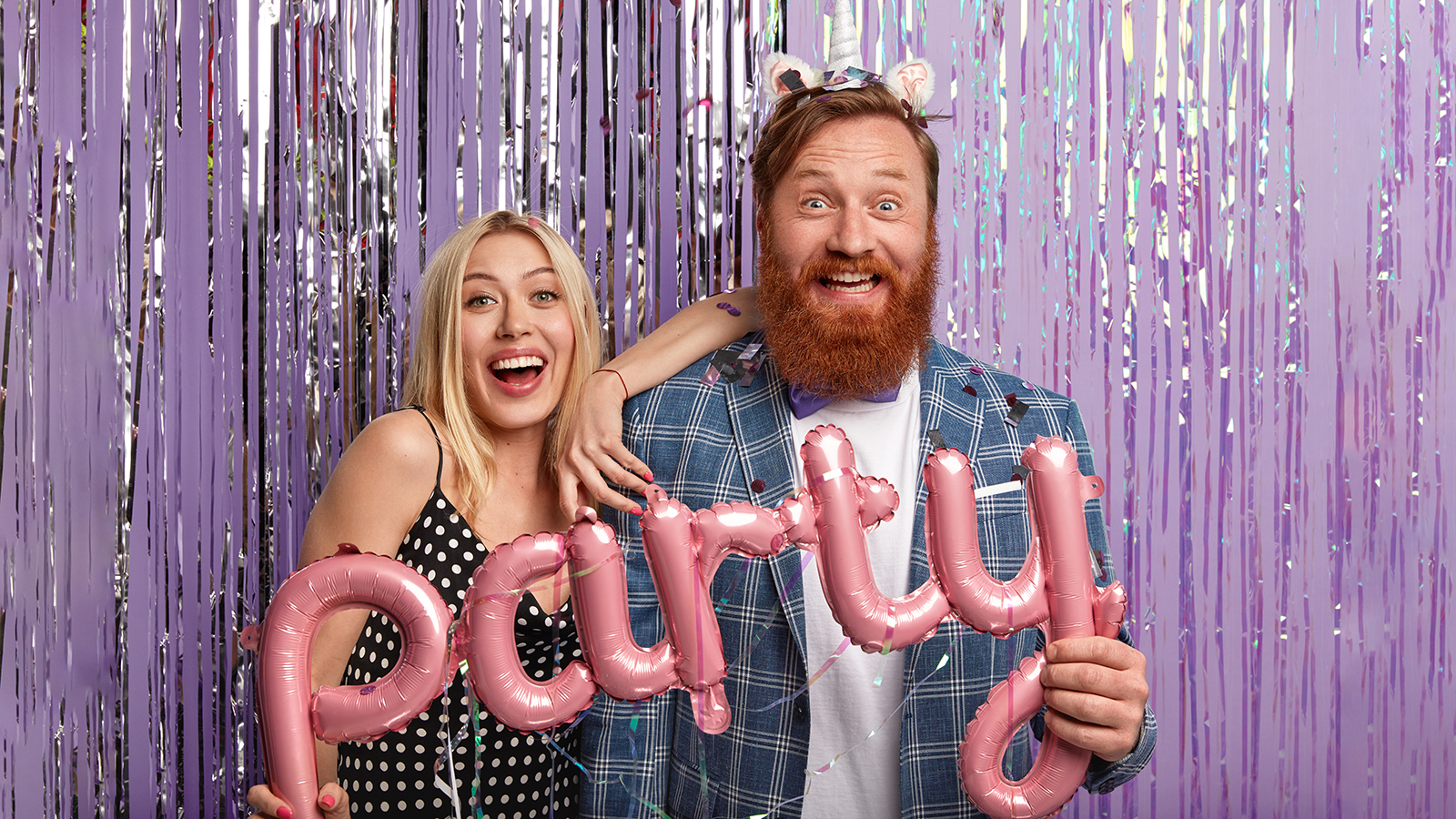 Joyous couple stand next to each other, have fun indoor, pose in decorated photozone, enjoys spare time, hold pink balloons in form of letters, have overjoyed facial expressions invite friends to join