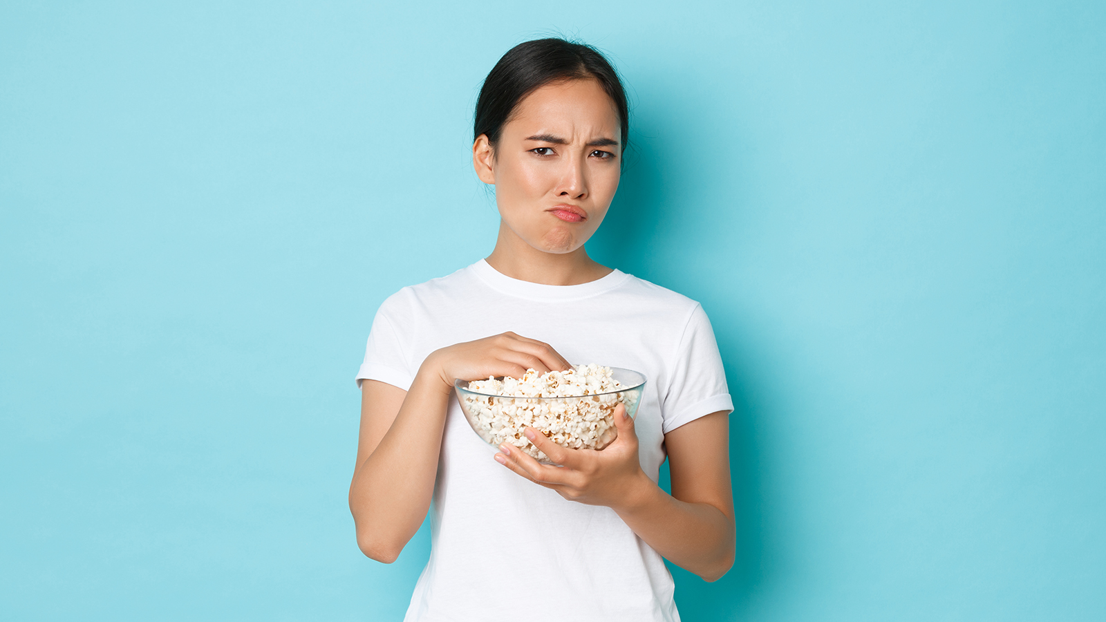Lifestyle, leisure and emotions concept. Skeptical and displeased asian cute girl watching movie and eating popcorn, grimacing from cringe content, looking suspicious, standing light blue background