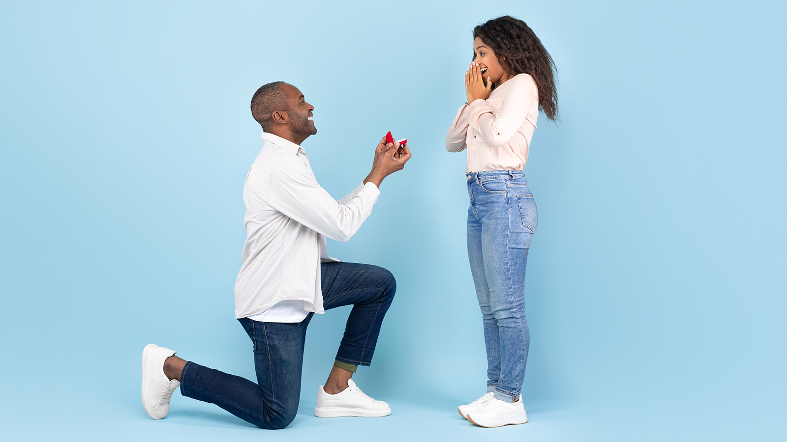 Marry me. Black middle aged man holding giving open box with engagement ring to excited young woman, asking her to be his wife during romantic date standing on one knee, blue studio background