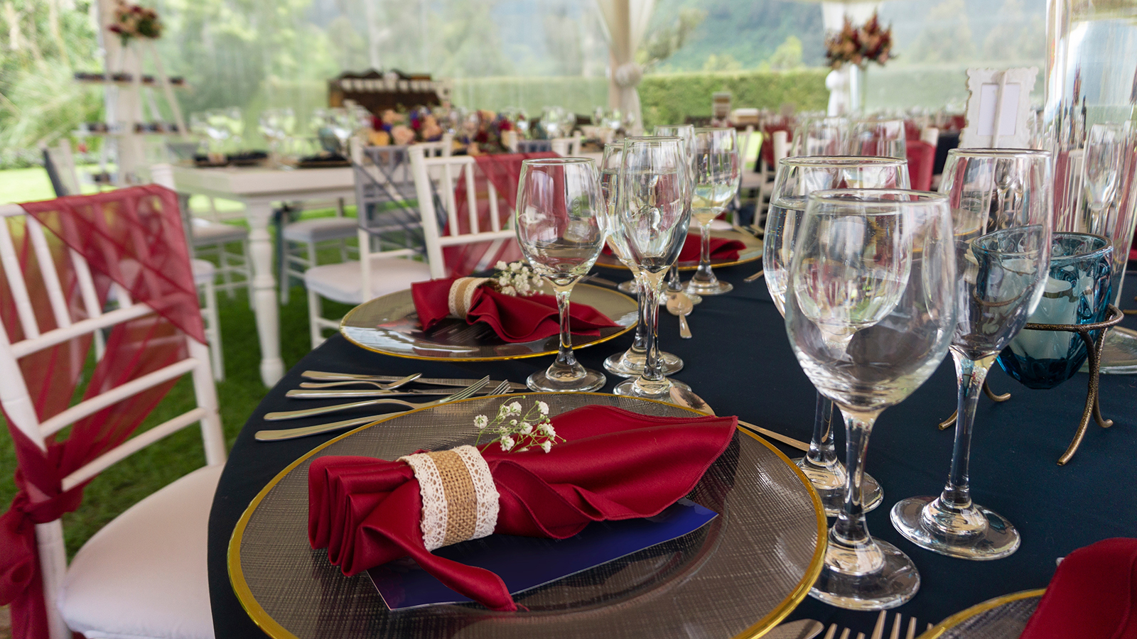 Party table in a garden, decorated with red napkins, blue tablecloth and crystal glasses with a background of white chairs