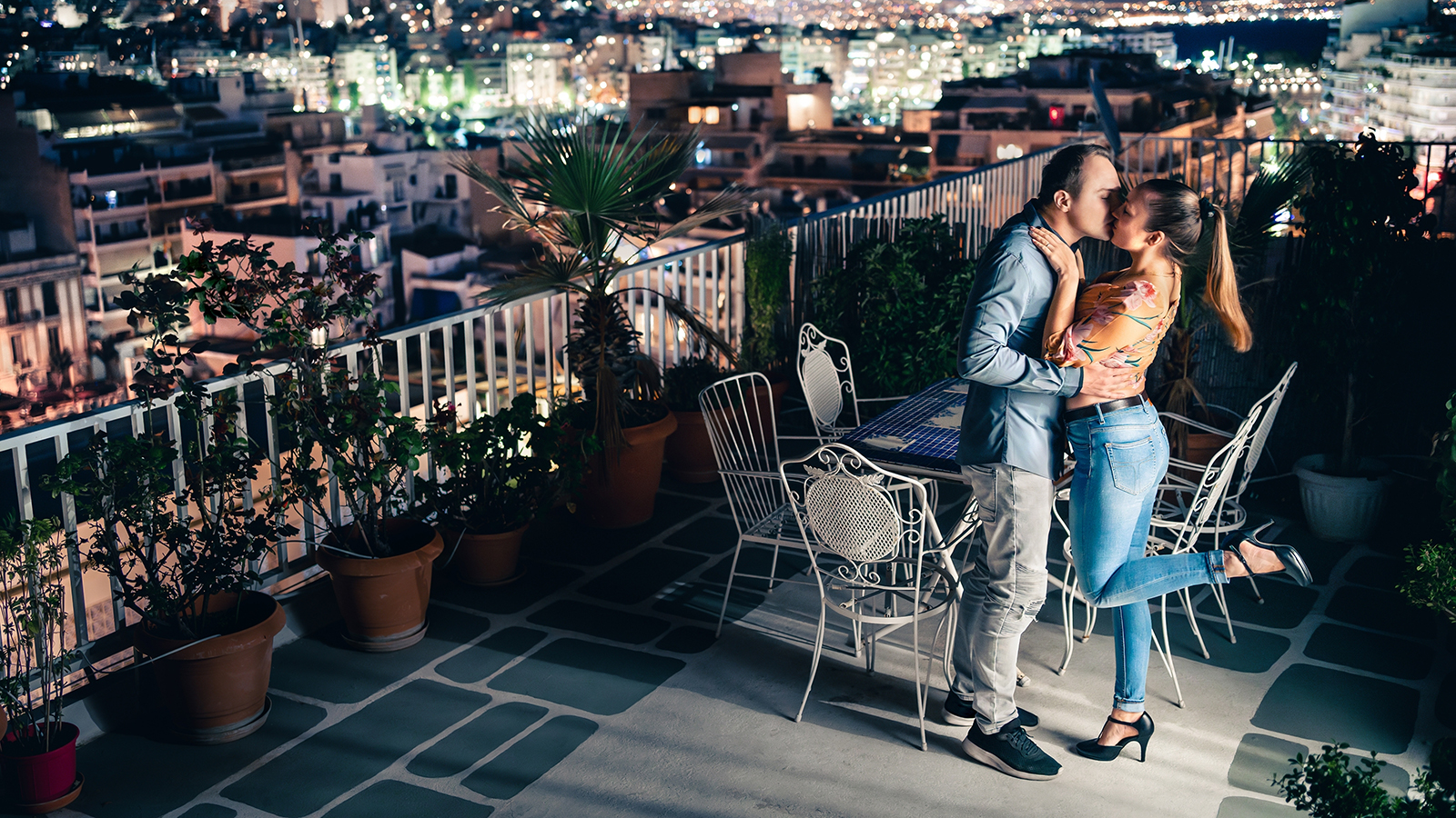 Couple at night, city lights view. Romantic date or proposal. Man and woman kiss and embrace on balcony or roof terrace. Dark blue sky and charming town in the background in summer. Happy relationship
