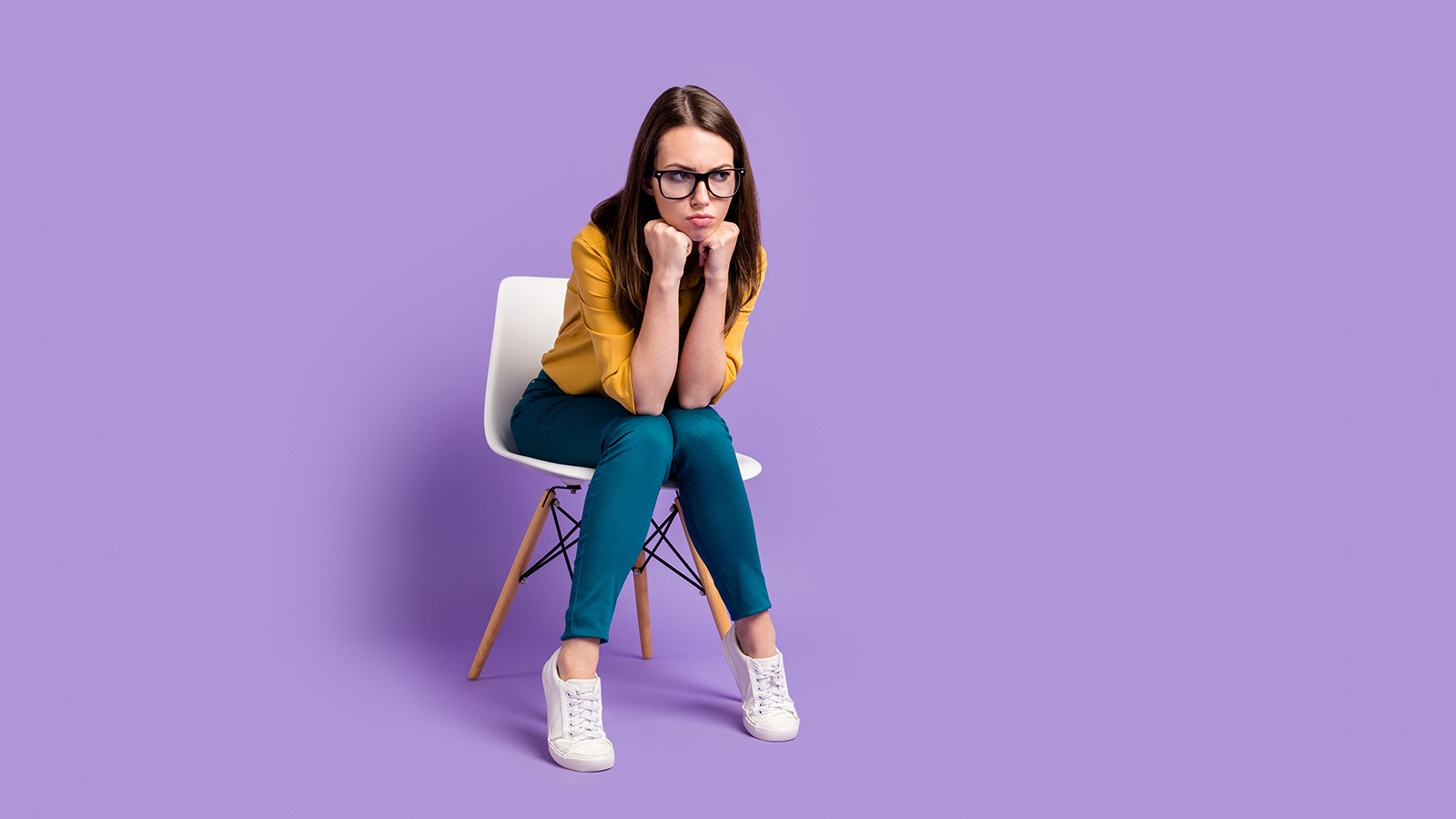 Full length photo of lady sit chair arms chin look empty space wear yellow shirt blue pants specs sneakers isolated purple color background