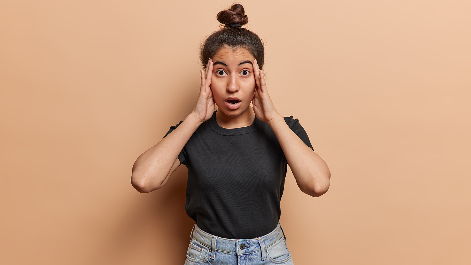 Concerned shocked Latin woman gasping with open mouth keeps hands on temples feels worried has big problems sees no way out wears black t shirt and jeans isolated over brown background. Omg concept