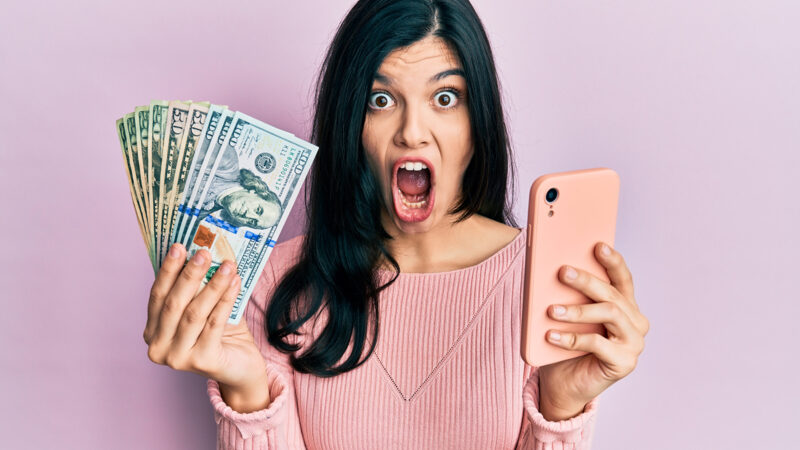 Young hispanic woman using smartphone holding united states dollar banknotes afraid and shocked with surprise and amazed expression, fear and excited face.