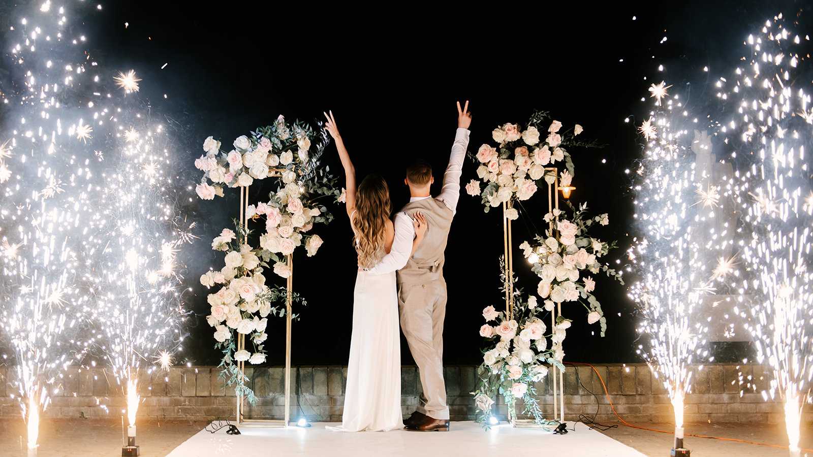 Young bride and groom stand near the wedding arch at night with lights, smoke and fireworks, silhouettes of the newlyweds