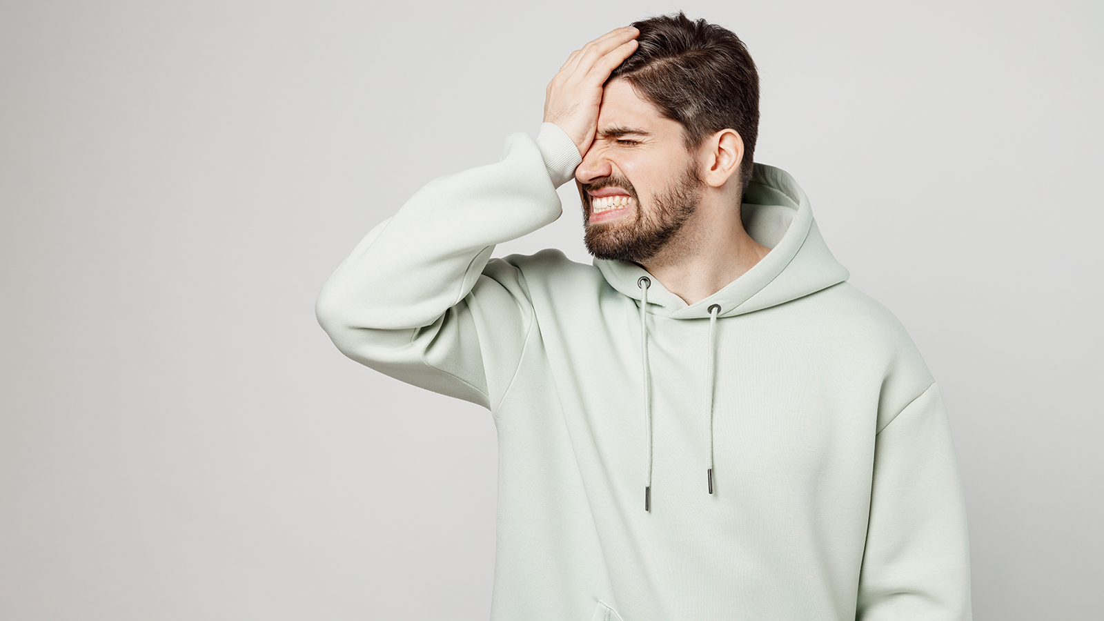 Young displeased sad unshaven caucasian man wear mint hoody put hand on face facepalm epic fail mistaken omg gesture isolated on plain solid white background studio portrait. People lifestyle concept