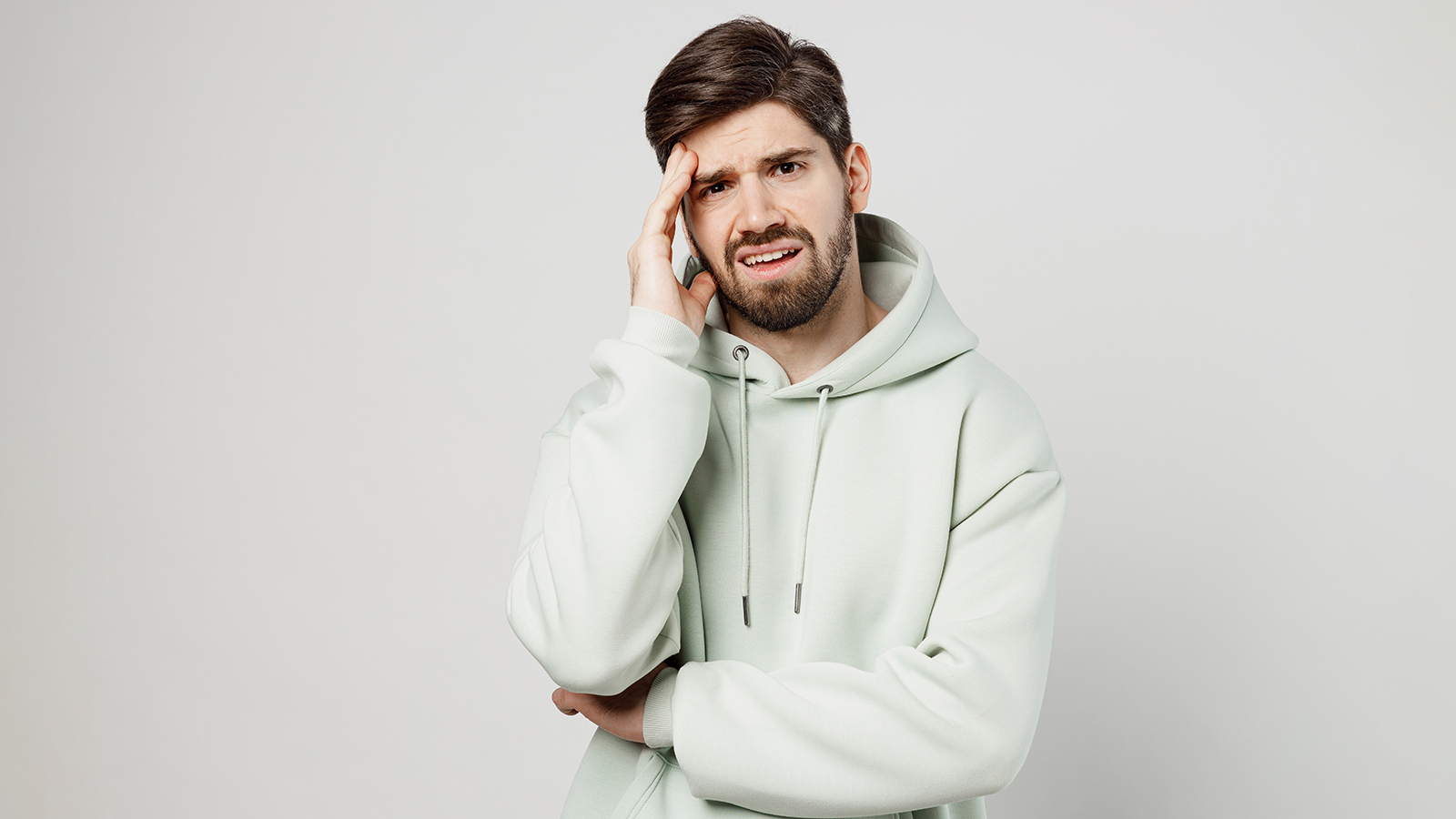Young dissatisfied sad mistaken depressed unshaven caucasian man wear mint hoody look camera prop up forehead head isolated on plain solid white background studio portrait. People lifestyle concept