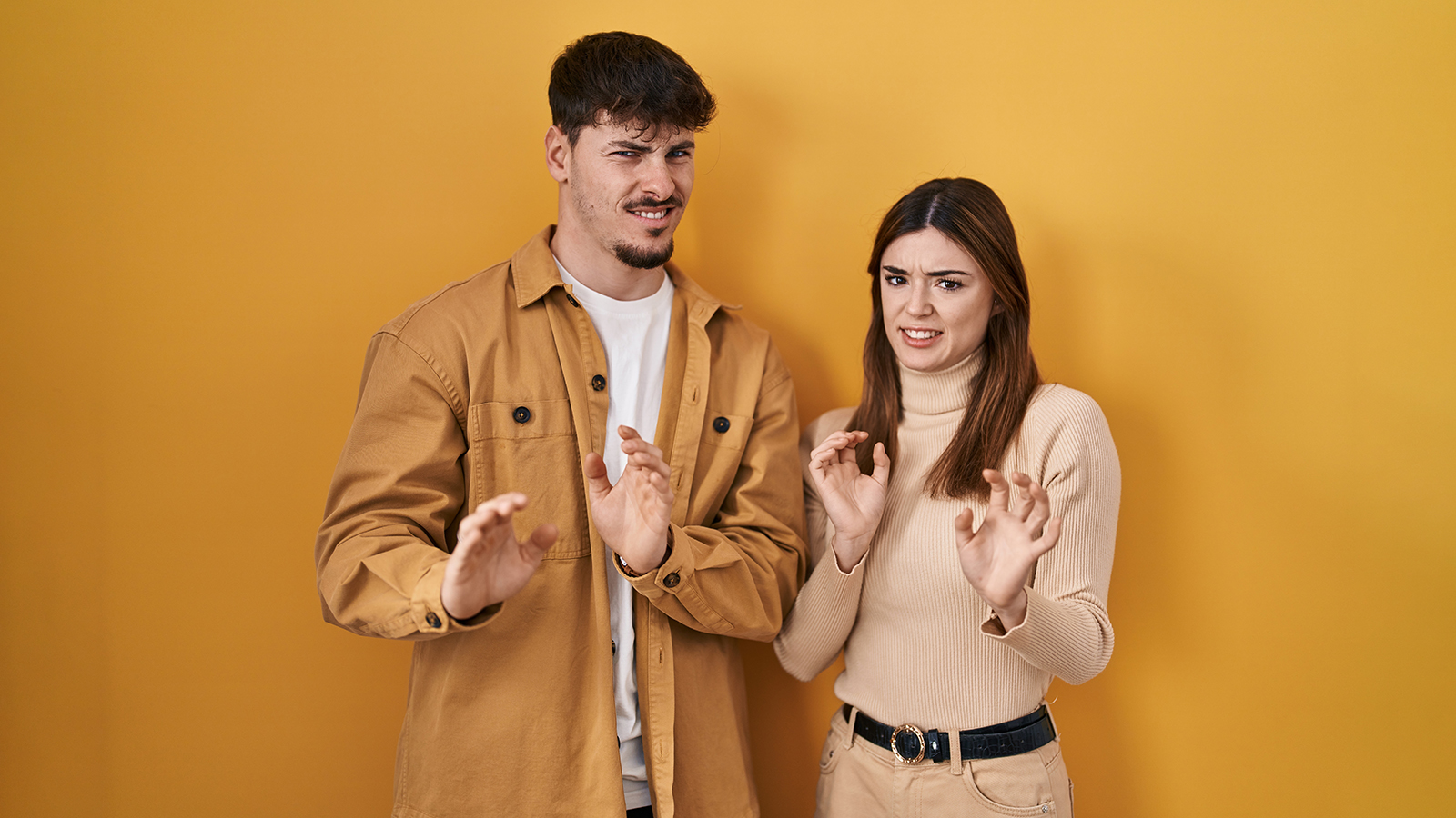 Young hispanic couple standing over yellow background disgusted expression, displeased and fearful doing disgust face because aversion reaction.