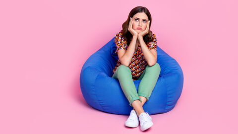 Full body length photo of young stressed sit bean bag unhappy minded bored woman touch cheekbones look empty space isolated on pink color background