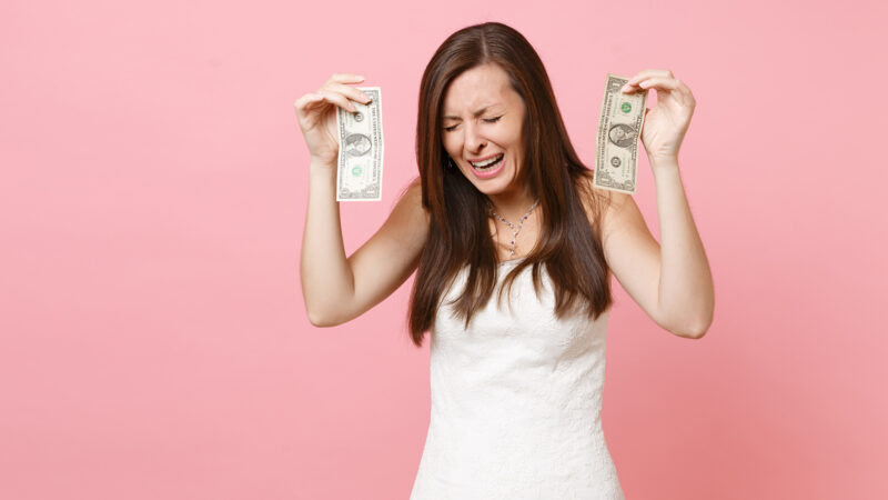 Portrait of upset sad bride woman in white wedding dress crying and holding one dollar bills isolated on pastel pink background. Lack of money. Organization of wedding celebration concept. Copy space