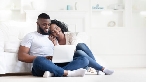 Planning summer vacation. Cute african american couple searching hotel and resort online, using laptop on floor at home, free space