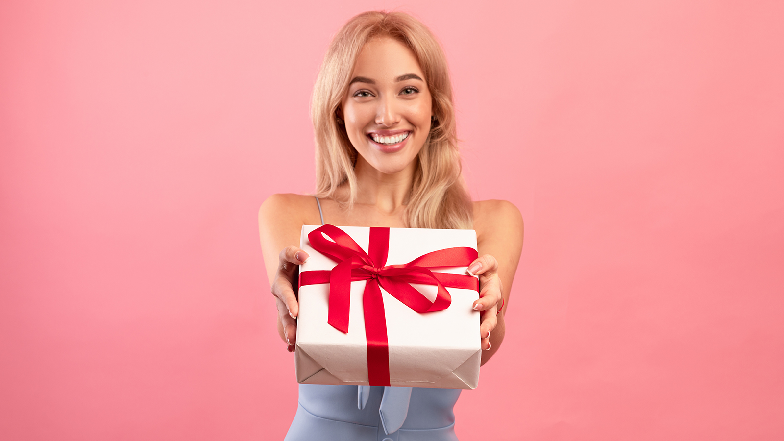 Lovely young woman giving wrapped gift box at camera over pink studio background. Cheerful millennial lady offering holiday present with smile, celebrating birthday or anniversary