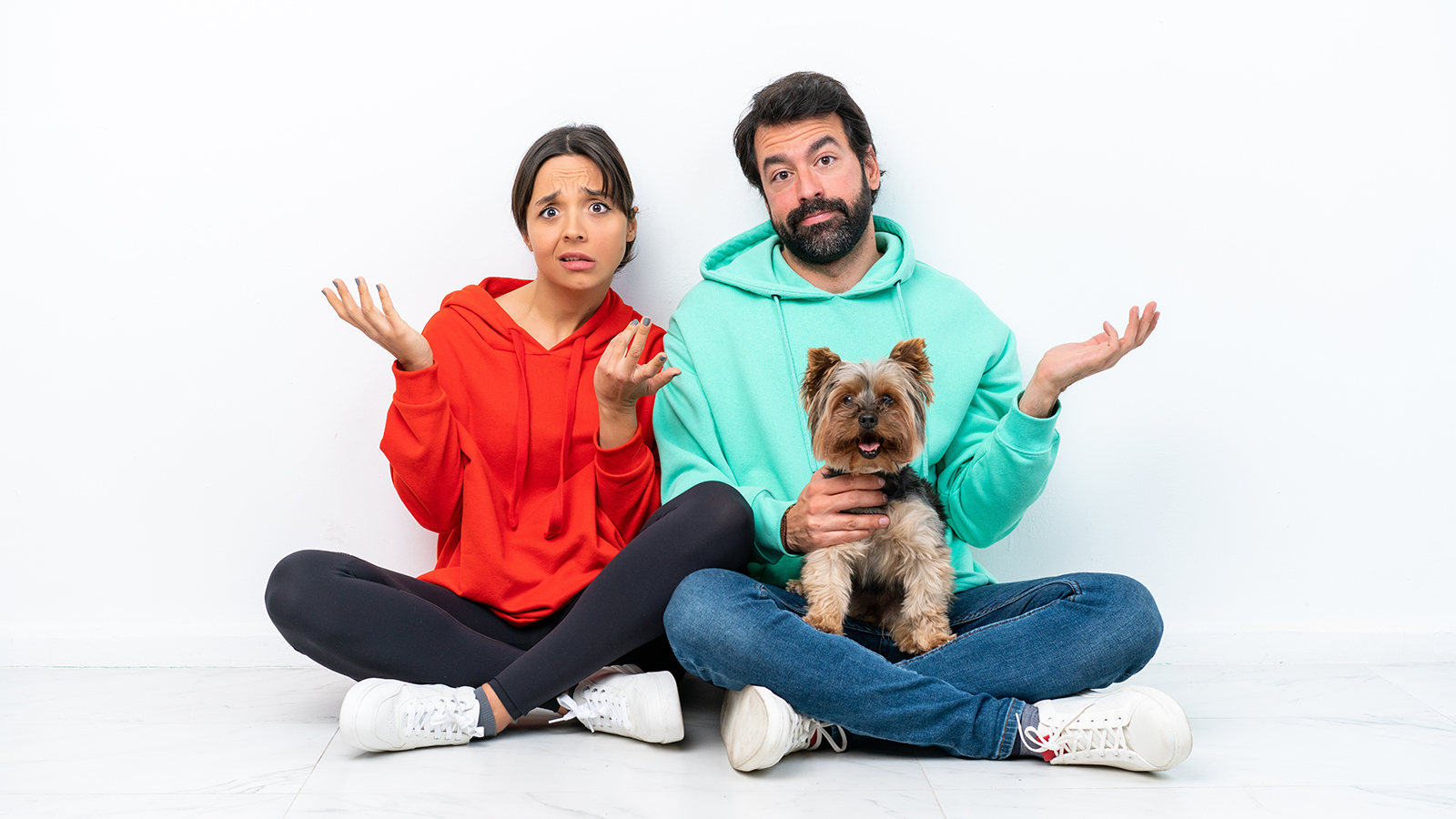 Young caucasian couple sitting on the floor with their pet isolated on white background having doubts and with confuse face expression