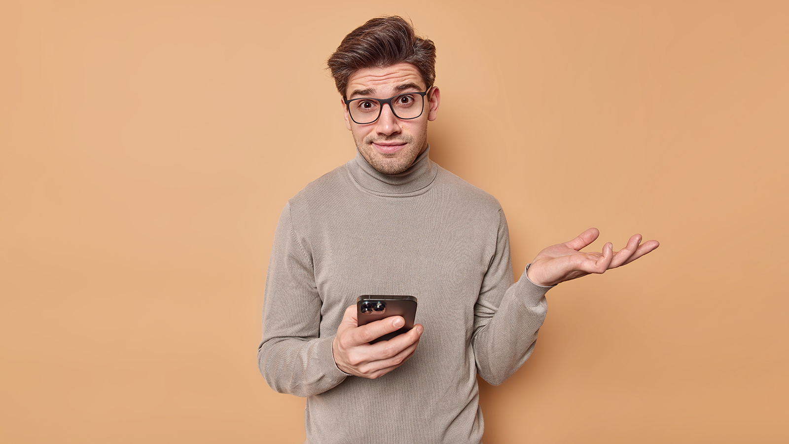 Confused questioned young man shrugs shoulders wears optical glasses and turtleneck holds mobile phone reads something strange browses newsfeed via social networks puzzled about online content