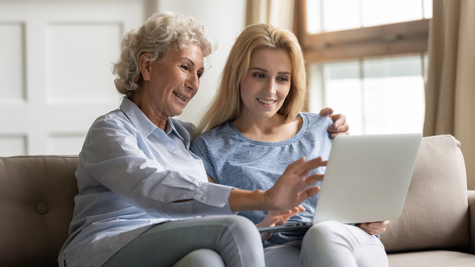 Happy older mature woman cuddling attractive grownup daughter, using computer at home. Smiling young lady teaching elderly mother use applications on laptop, shopping online, sitting together on sofa.