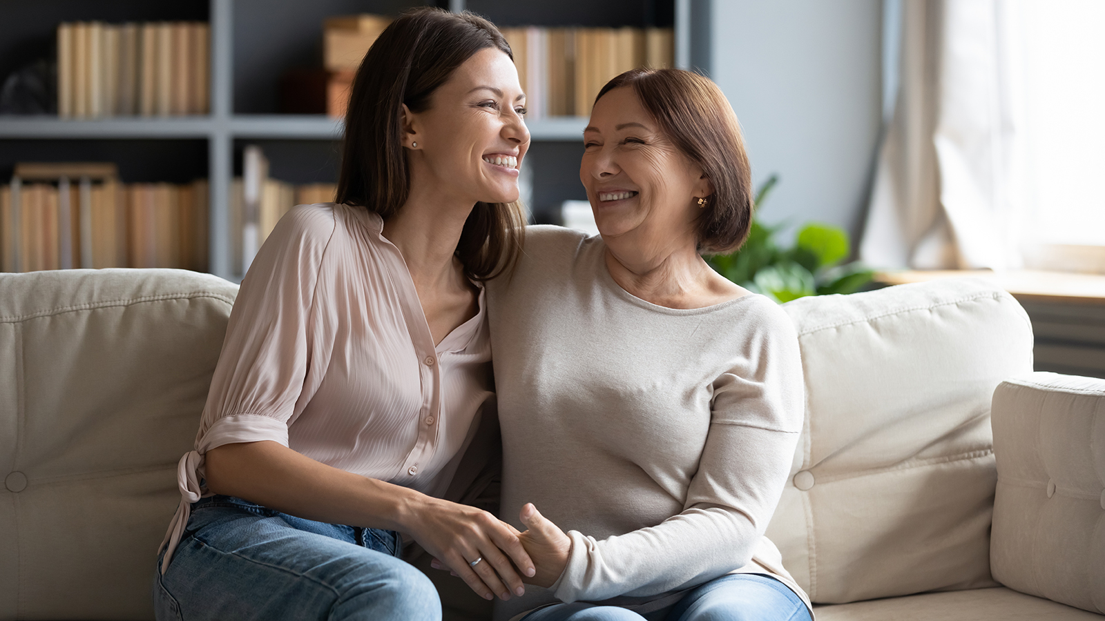 Overjoyed young woman with mature mother chatting, laughing at joke, enjoying pleasant conversation, middle aged mum and grownup daughter hugging, having fun, sitting on cozy couch at home