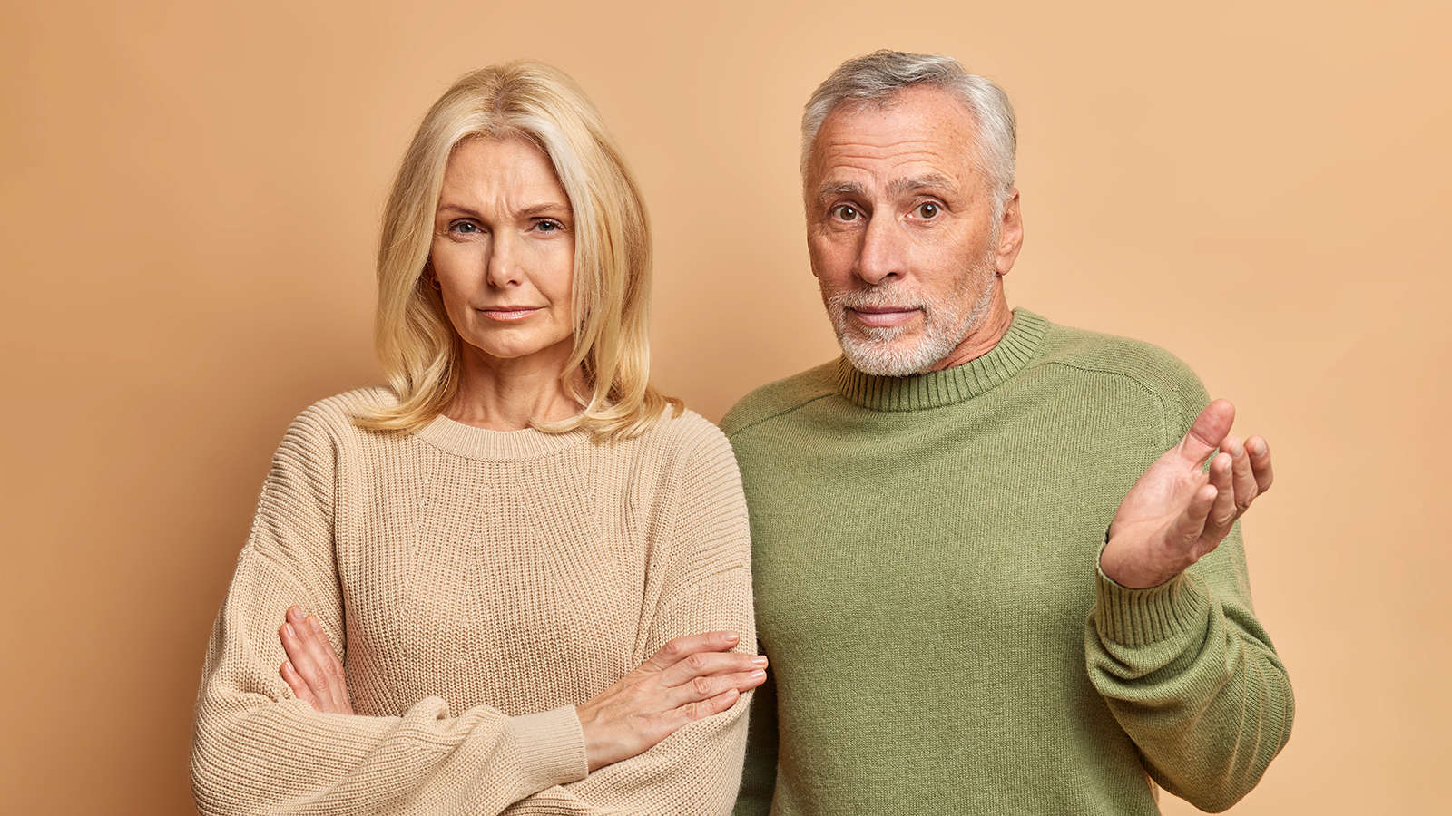 Self assured blonde middle aged woman stands with arms folded and looks seriously at camera her grey haired bearded husband shrugs shoulders in confusement being unaware. Family spouses indoor