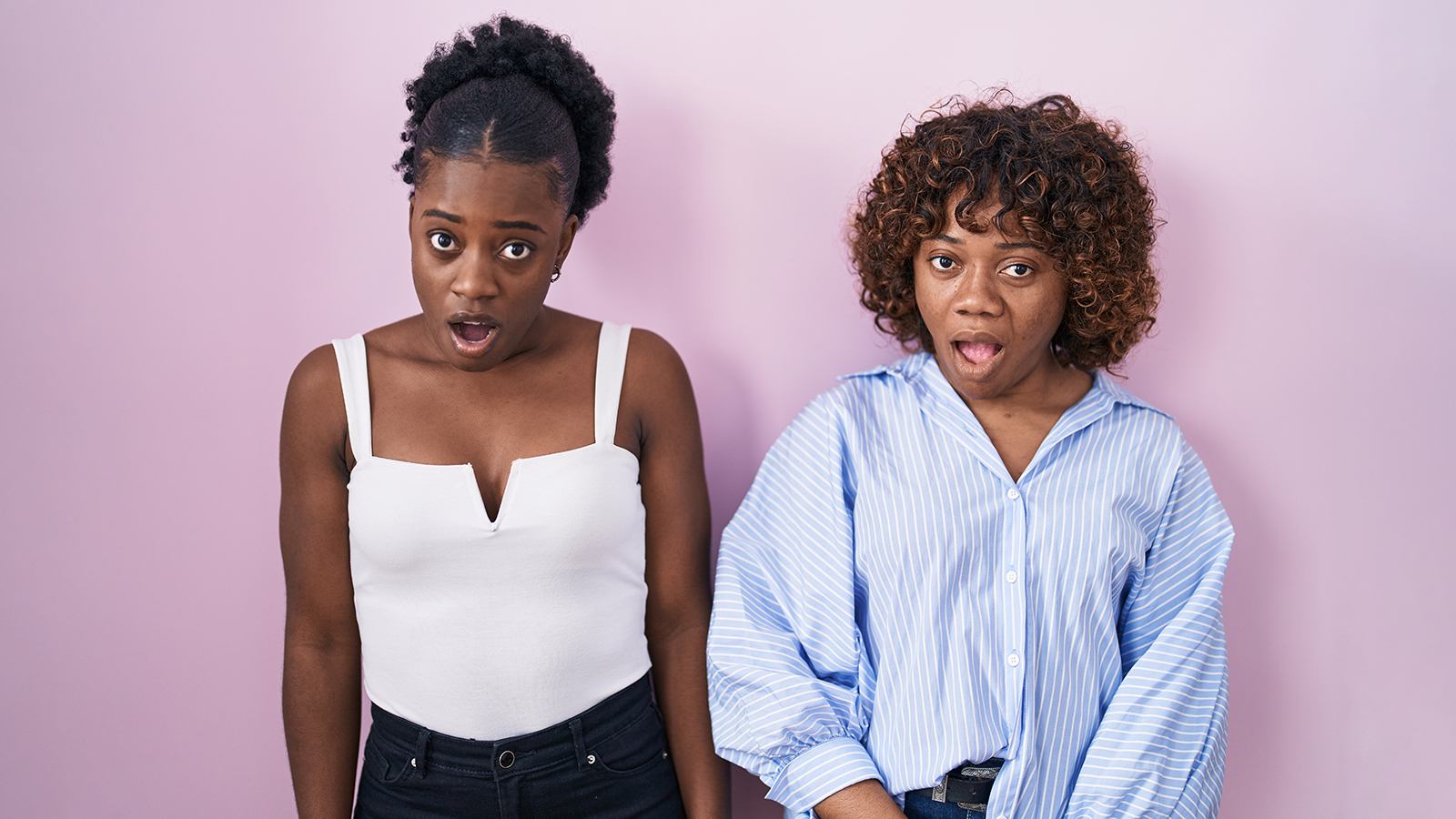 Two african women standing over pink background in shock face, looking skeptical and sarcastic, surprised with open mouth