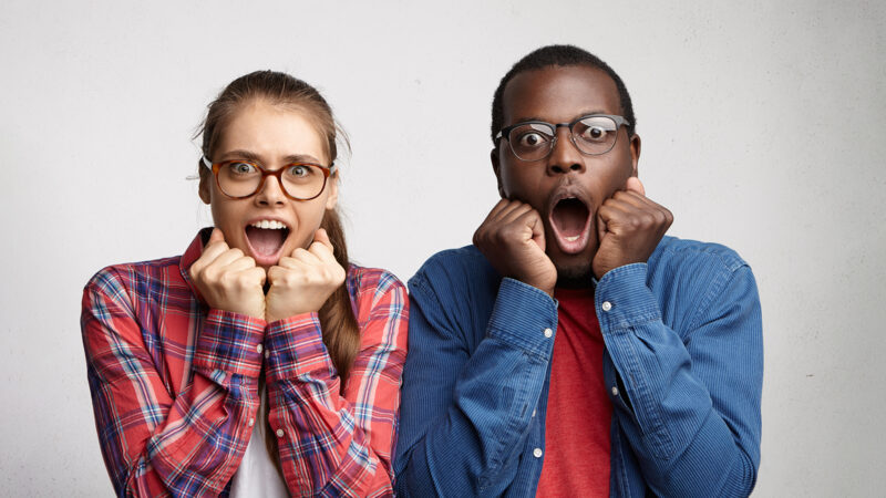Terrified and puzzled couple with opened mouths holding fists under their chins. Full face portrait of two interracial friends being shocked by bad news. International couple in panic and terror