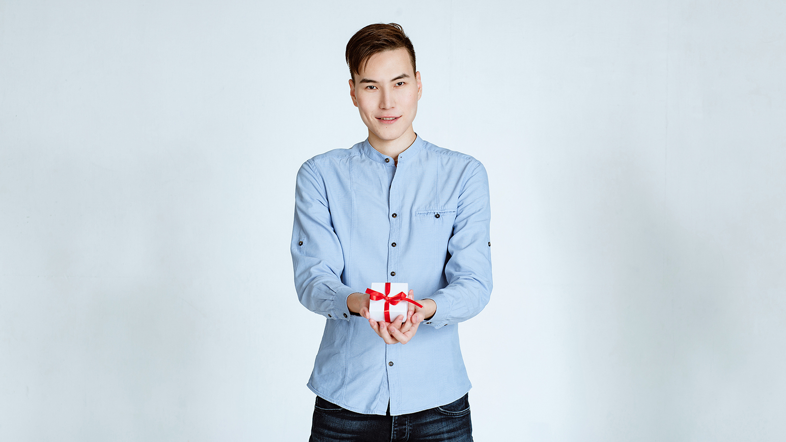 young handsome male giving a gift, looks in the camera and smiles. the young man of Asian with a fashionable stylish hairstyle and in a blue shirt holds in hand a box with a gift with a red tape