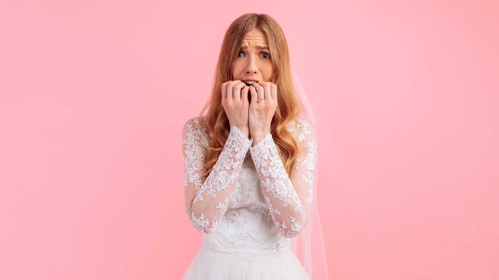A frightened bride in a wedding dress and with a bouquet of flowers, holding her hands to her mouth in fear on a pink background