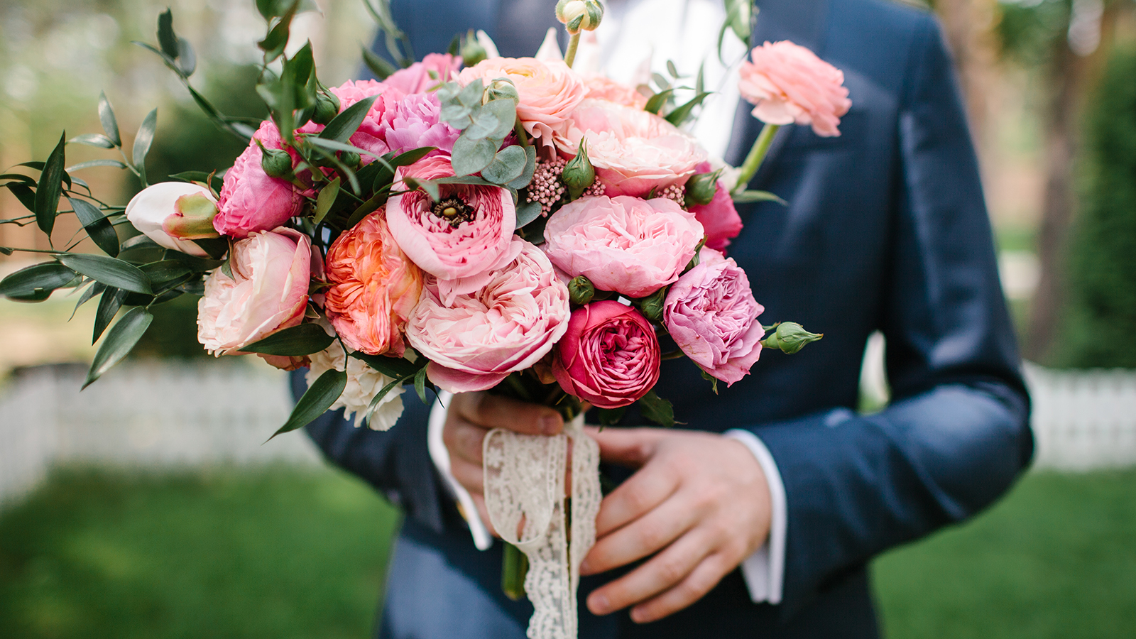a wedding bouquet of pink Ranunculus, red roses, peach peonies, eucalyptus in groom hands in suit