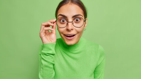 Photo of surprised cheerful woman with dark hair looks wondered through spectacles cannot believe her eyes dressed in casual jumper isolated over vivid green background. Human reactions concept