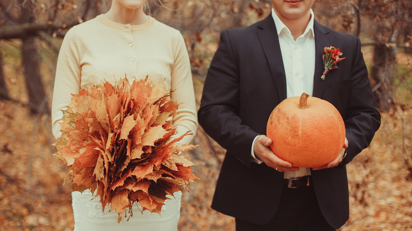 Autumn wedding. The groom is holding an orange pumpkin, the bride is holding a bouquet of autumn leaves. Foliage with pumpkin. High quality photo. Thanksgiving or Halloween concept banner or postcard.
