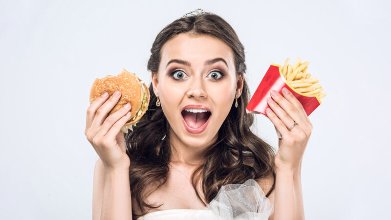 close-up portrait of shocked young bride in wedding dress with burger and french fries looking at camera isolated on white