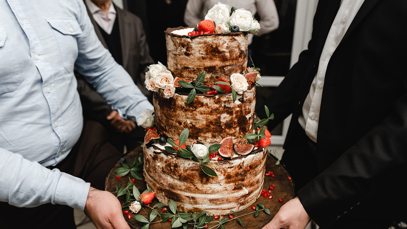 Elegant wedding cake decorated with roses and fruits with bride and groom on background. Wedding ceremony. Beautiful pie on the table. Brides cut cake together. Close up