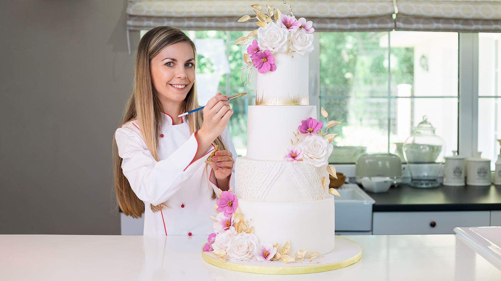 confectioner in chefs uniform standing in the modern kitchen, holding a brush and gold paint, decorating a four tier white, pink and gold wedding cake
