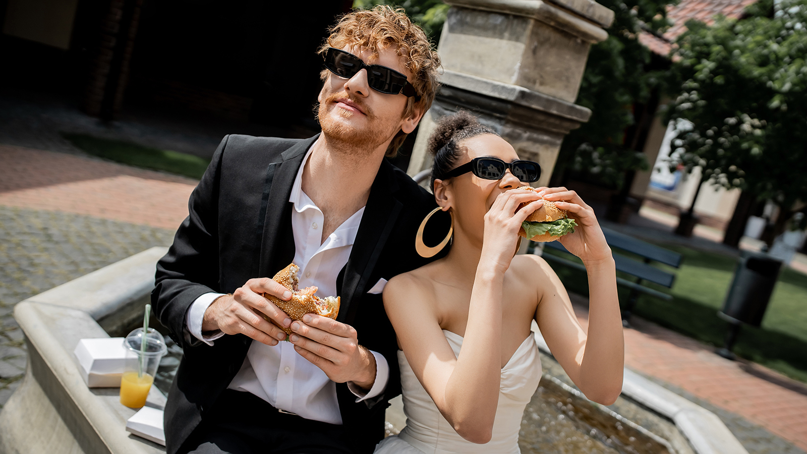 african american bride eating burger near redhead groom in sunglasses near fountain, outdoor wedding