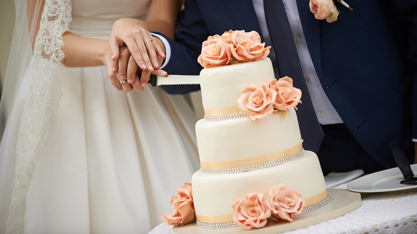 bride and groom cut the wedding cake. The cake is decorated with beige and peach-colored roses. groom is dressed in blue wedding suit and bride in a white wedding dress.