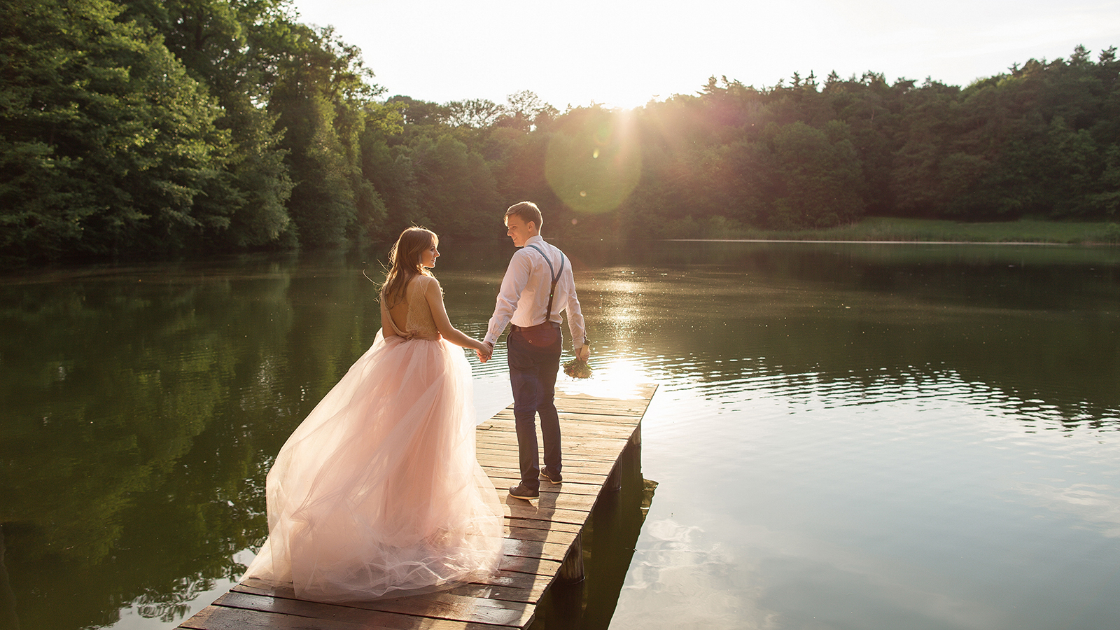 Wedding couple walking on bridge near lake on sunset at wedding day. Bride and groom in love