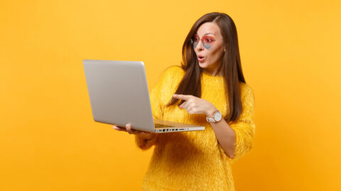 Surprised young woman in sweater heart eyeglasses working pointing index finger on laptop pc computer isolated on bright yellow background. People sincere emotions lifestyle concept. Advertising area